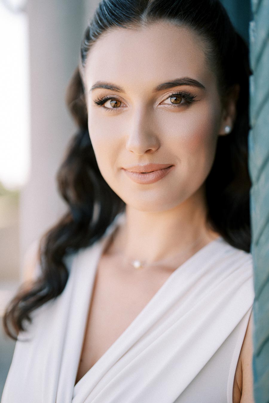 A woman with long dark hair and a subtle smile, wearing a white top, stands against a blurred background, showcasing natural beauty and elegance.