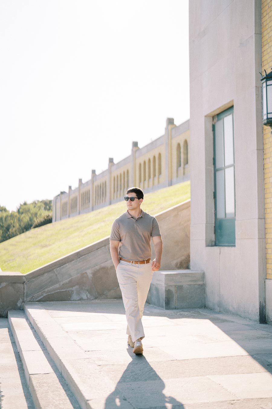 A person wearing sunglasses and casual attire walks confidently outside a modern building with large windows and a backdrop of green grass and clear skies.