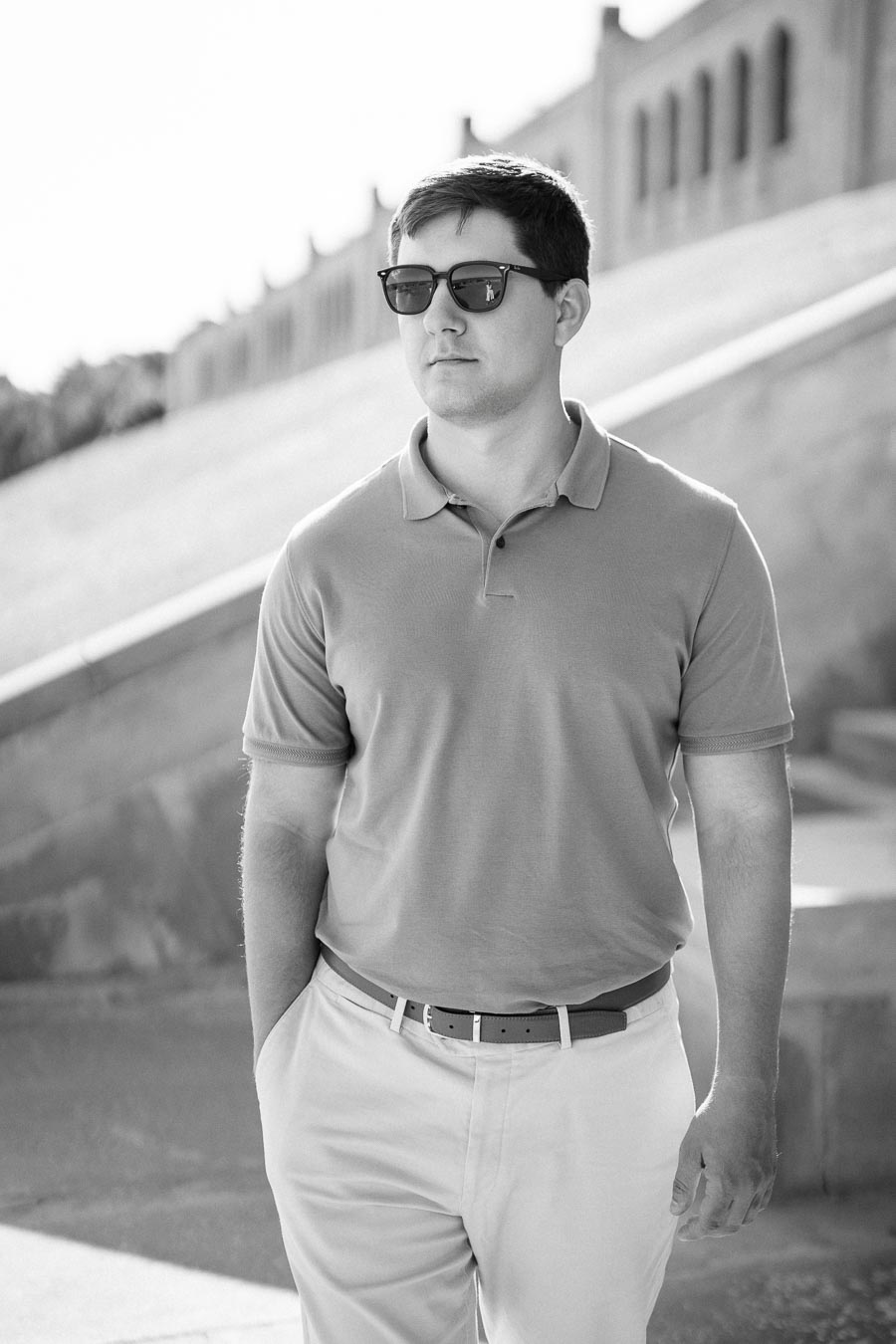 Black and white photo of a man wearing sunglasses and a polo shirt, walking confidently near a stone staircase.