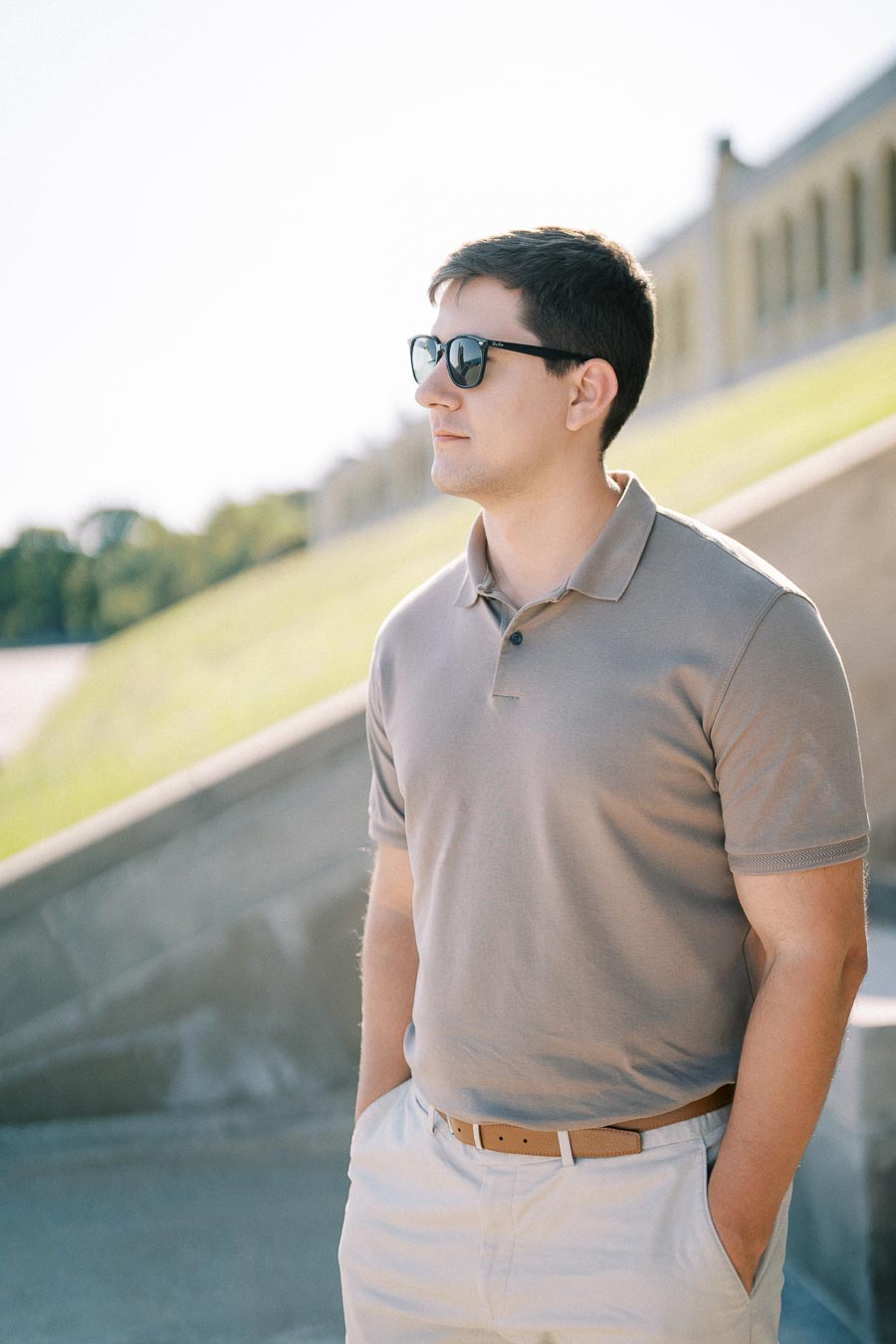 Young man in sunglasses wearing a gray polo shirt outdoors, standing with hands in pockets, against a bright, sunny background.