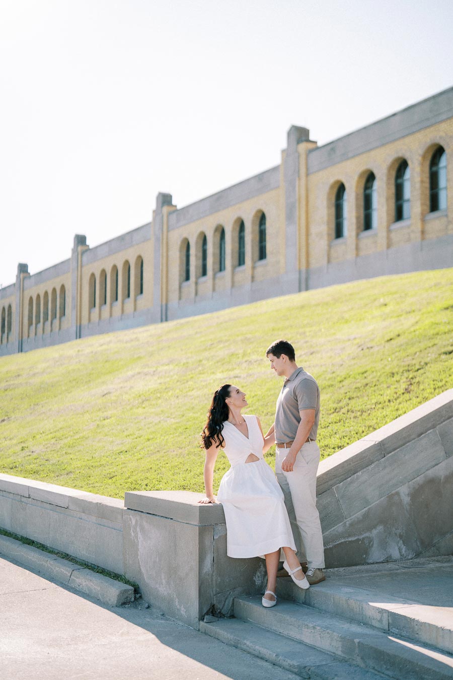 A couple sharing a romantic moment on stone steps in front of a historical building with arched windows, under a clear blue sky.