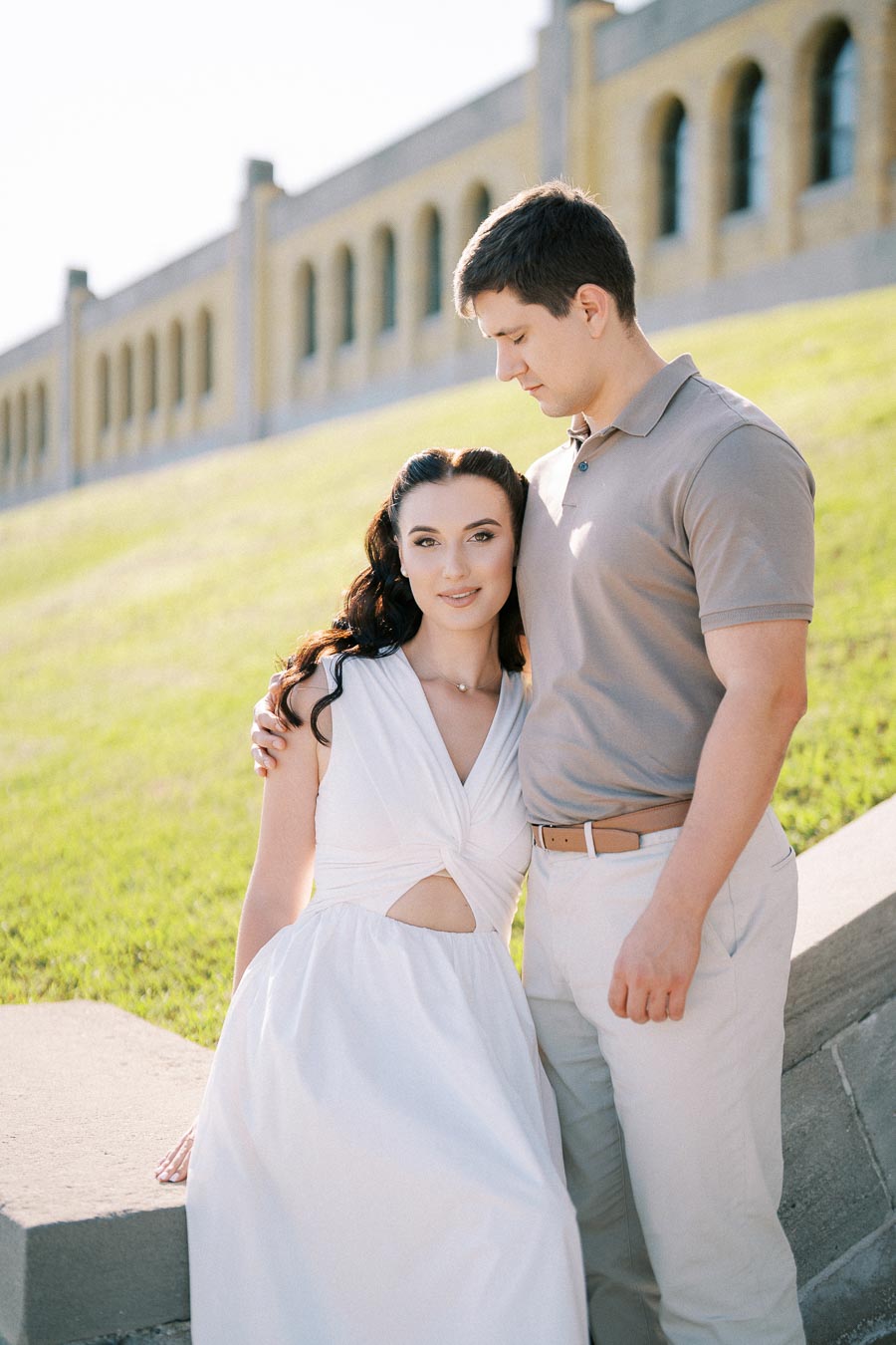 Couple posing affectionately in front of a historic building, woman in white dress and man in casual attire, sunny outdoor setting