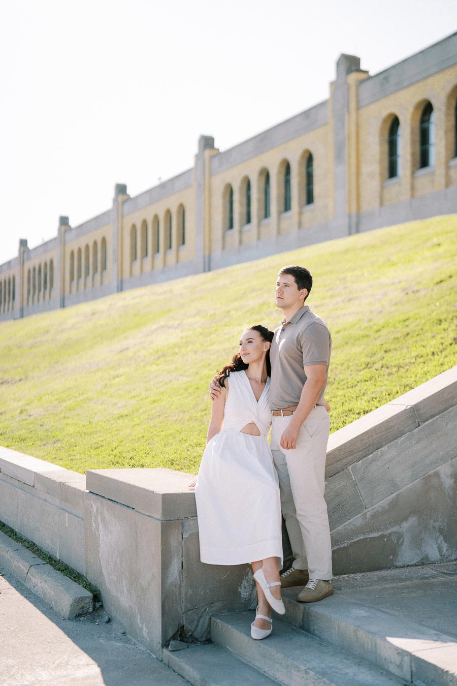 Couple posing on stone steps with historic building and grassy hill in background on a sunny day.