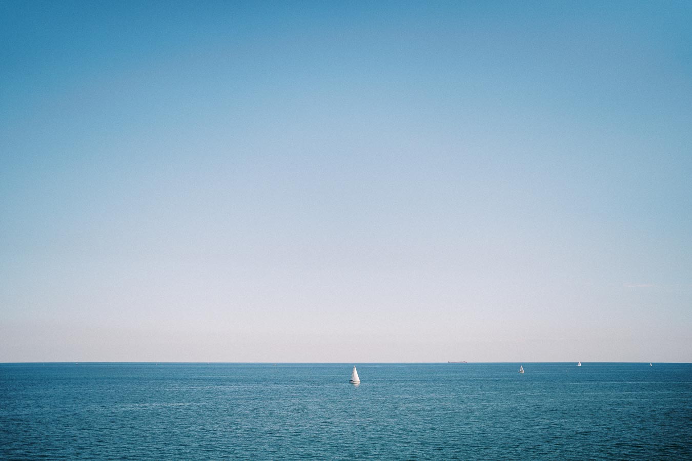 Calm ocean scene with a single sailboat on the horizon under a clear blue sky.