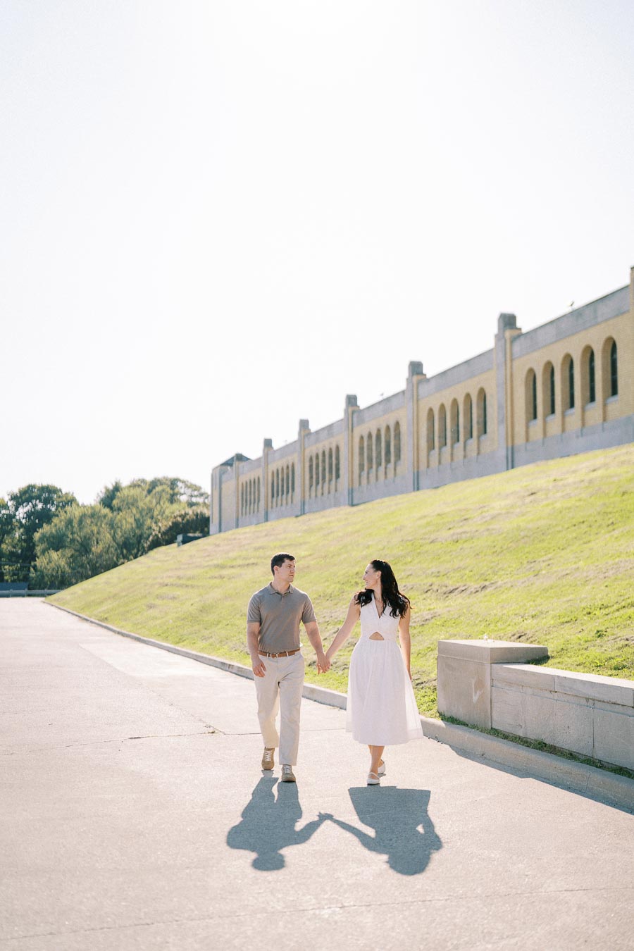 Couple holding hands and walking outside near a historic building on a sunny day, with green grass and trees in the background.