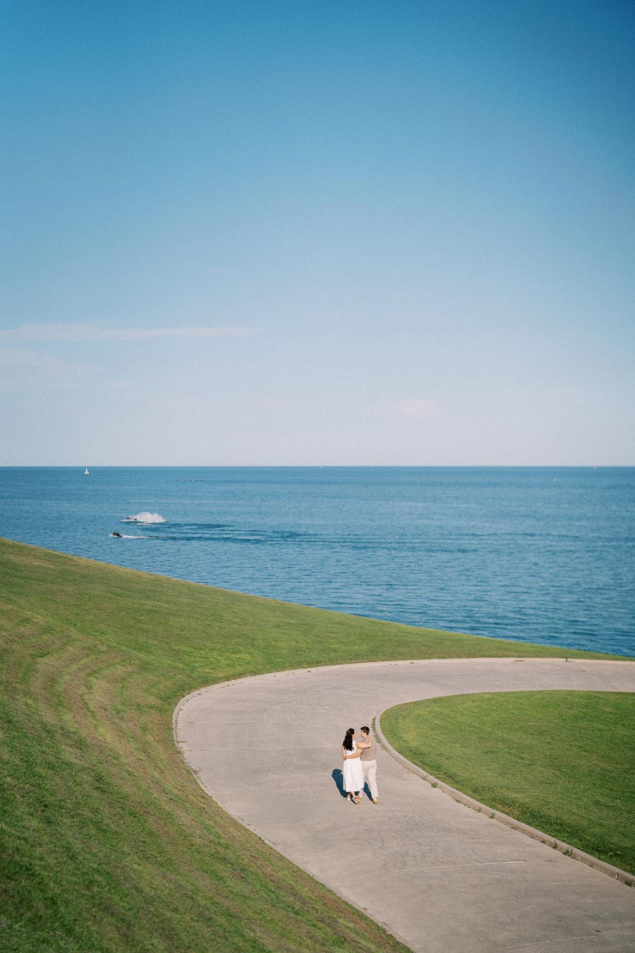 A couple walking on a curved path through a lush green park with a clear blue sky and a serene ocean in the background.