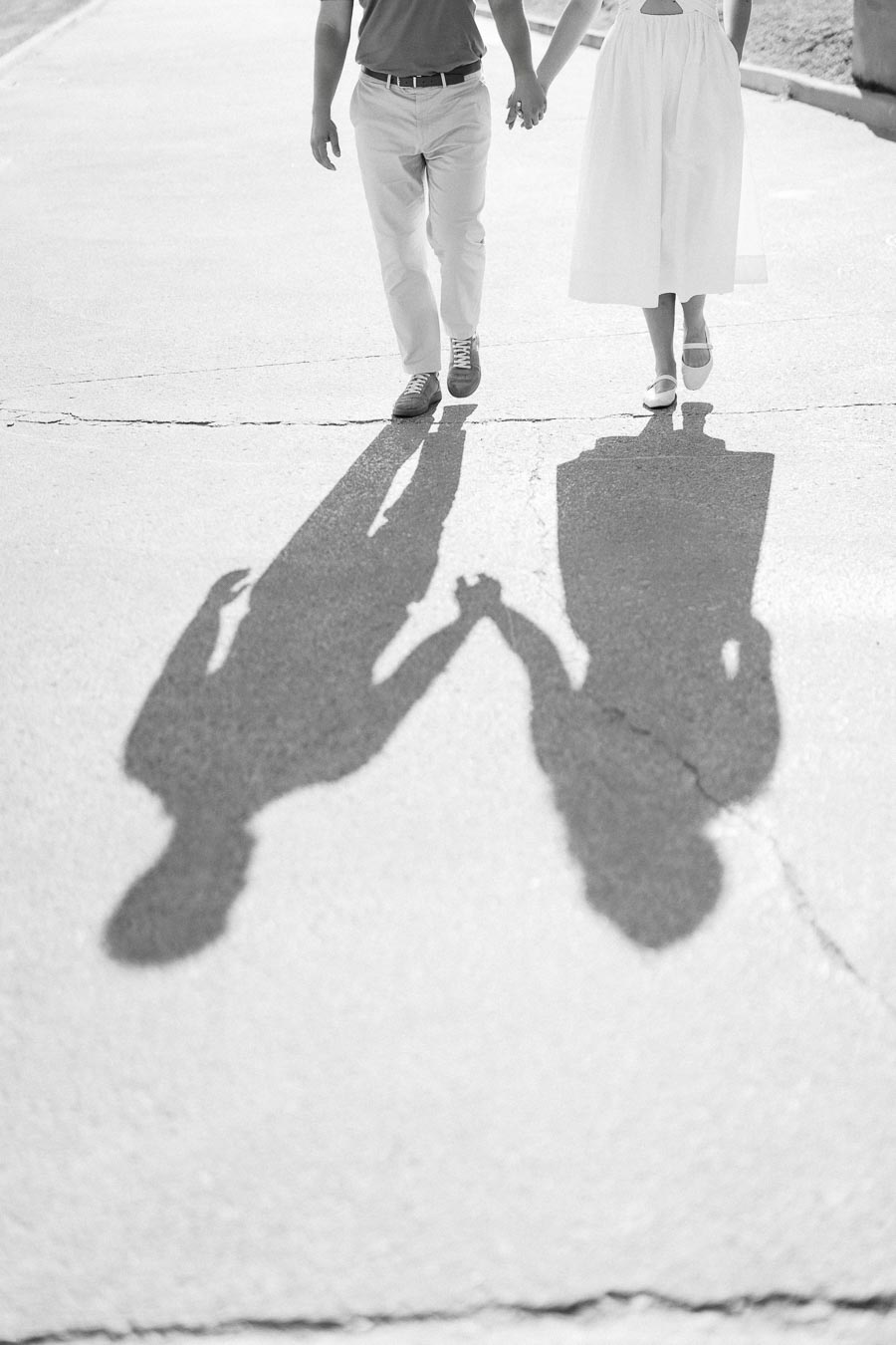 Black and white photo of a couple walking hand in hand, with their shadows cast on a sunlit pavement, symbolizing love and togetherness.