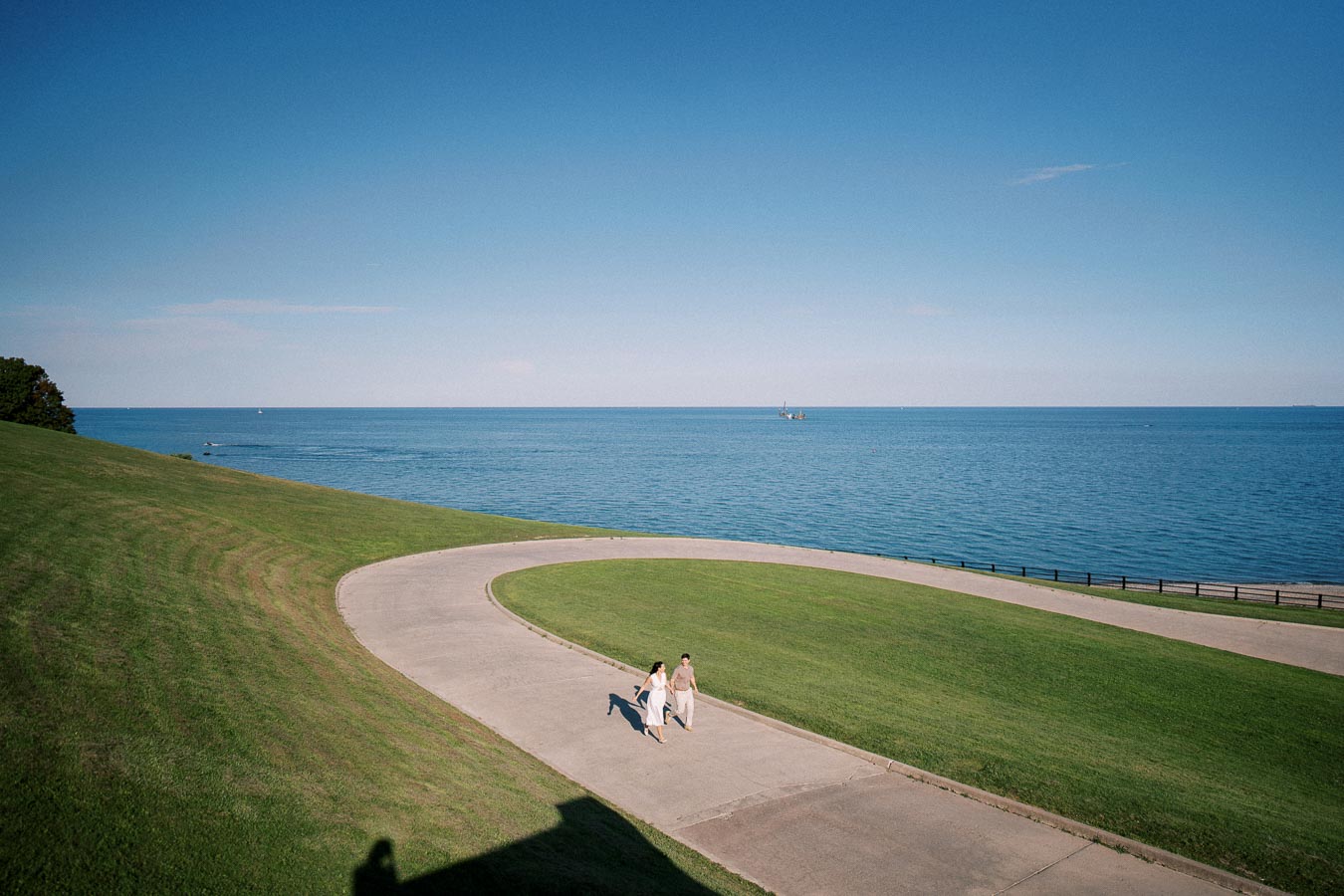 A couple walks on a curved path overlooking a serene blue lake on a sunny day, surrounded by lush green grass, with a clear sky in the background.