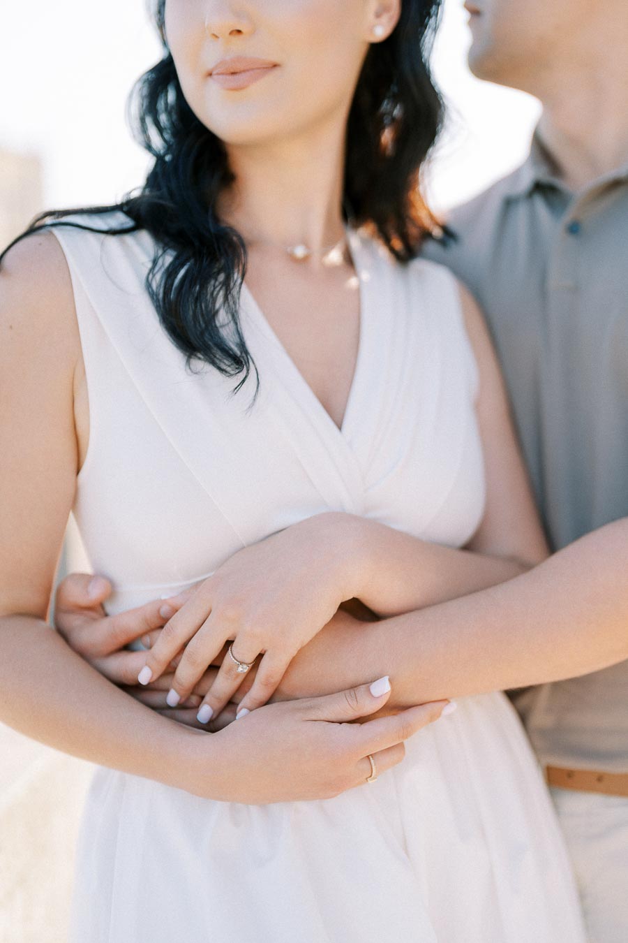 A couple embracing with hands clasped, highlighting an engagement ring on the woman's finger, outdoor sunny setting, symbolizing love and commitment.