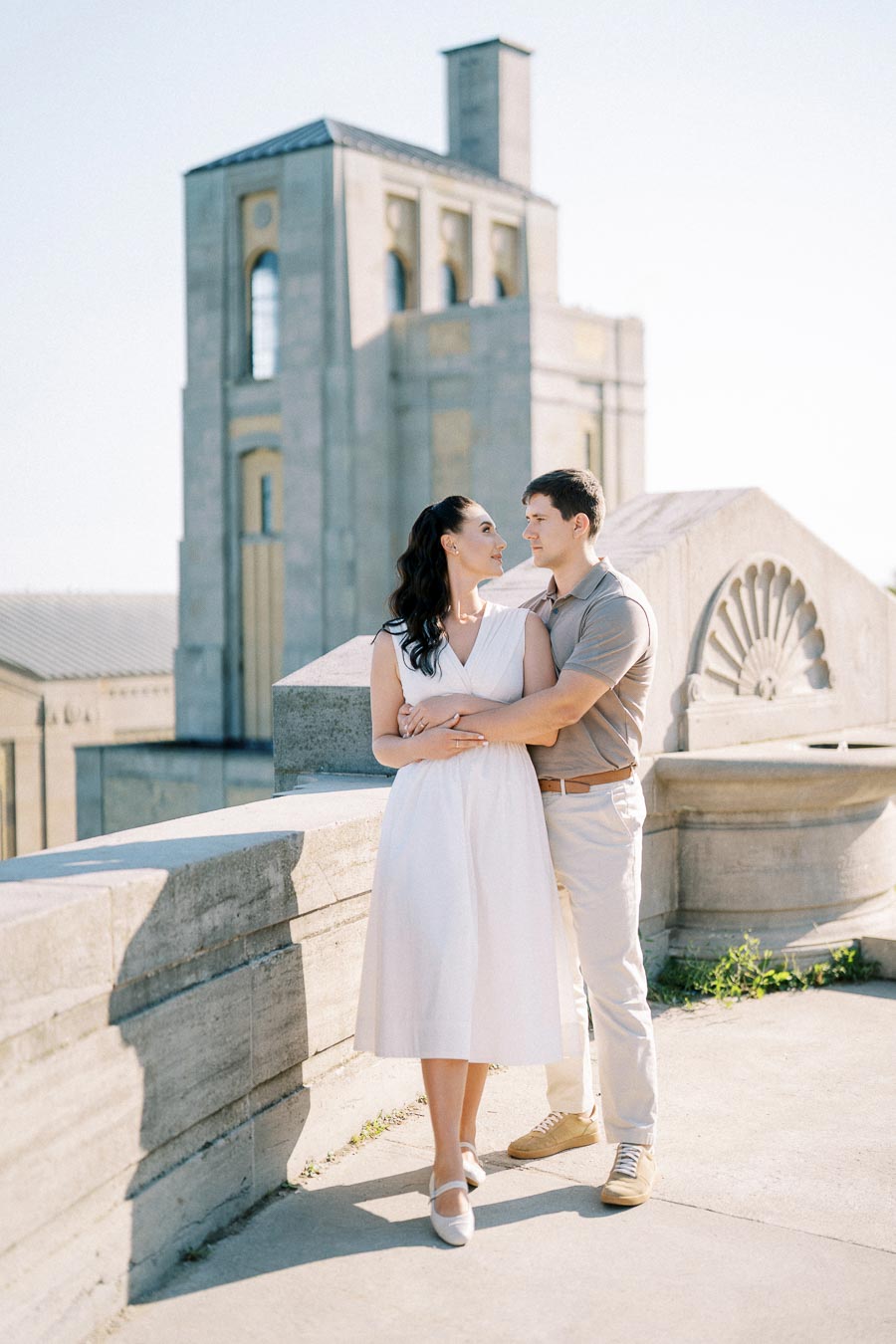 A couple embracing on a sunlit terrace with historic architecture in the background.