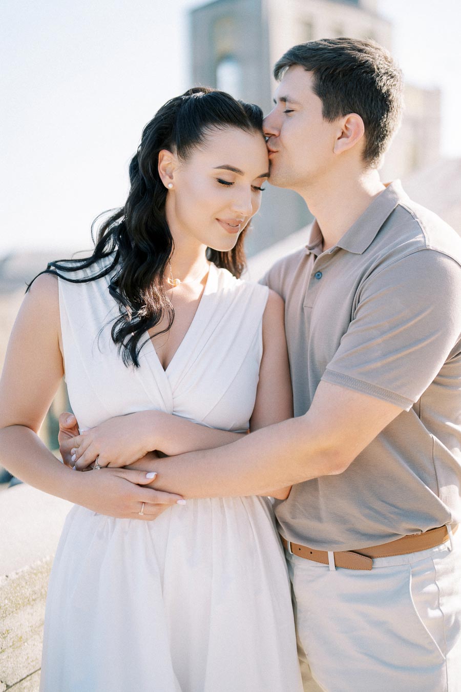 Affectionate couple embracing on a sunny day, with the man gently kissing the woman's forehead, conveying love and tenderness, outdoors with a blurred building in the background.