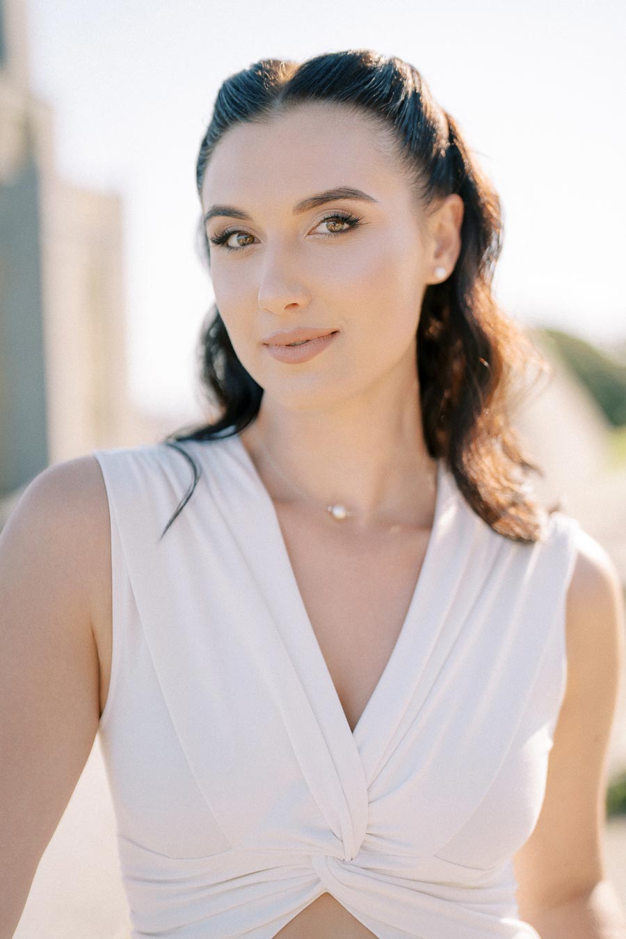 Portrait of a woman with dark hair styled in soft waves, wearing a white sleeveless top, standing outdoors against a blurred background, showcasing natural beauty and grace.