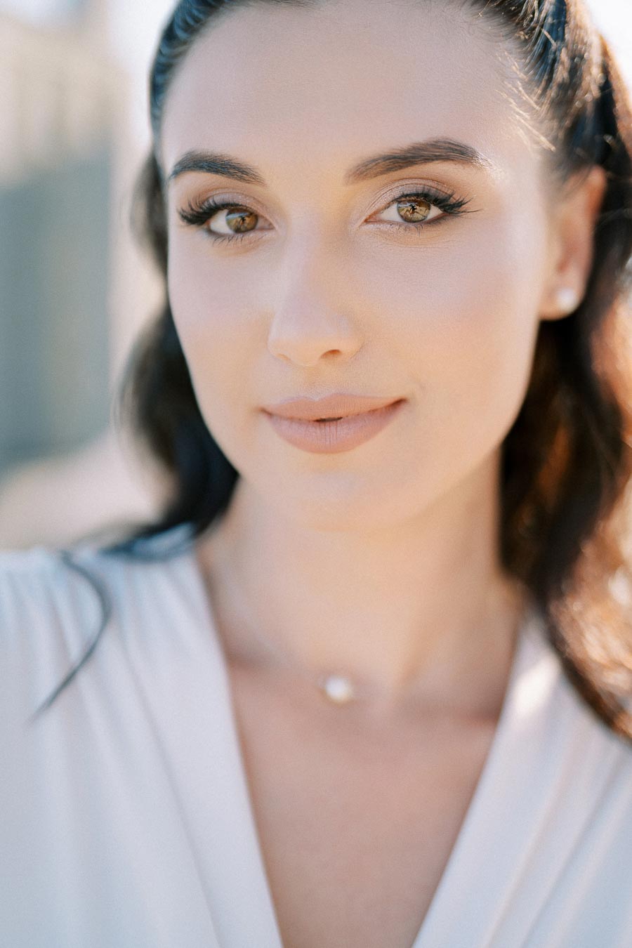 Close-up portrait of a woman with natural makeup and long dark hair, outdoors with blurred background.