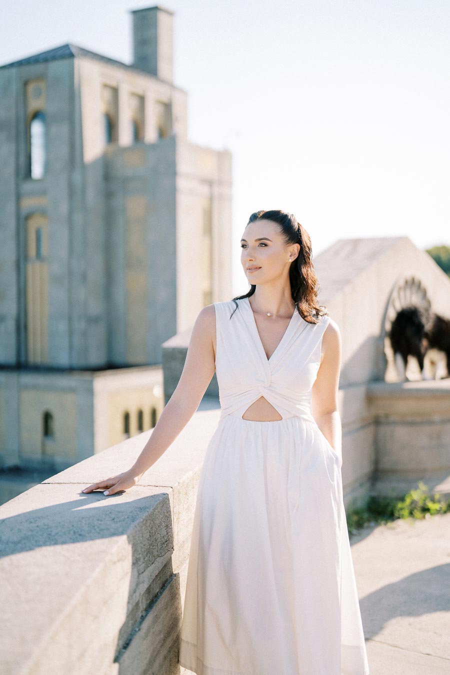 A woman in a white dress standing on a sunlit stone balcony with a historic building in the background.