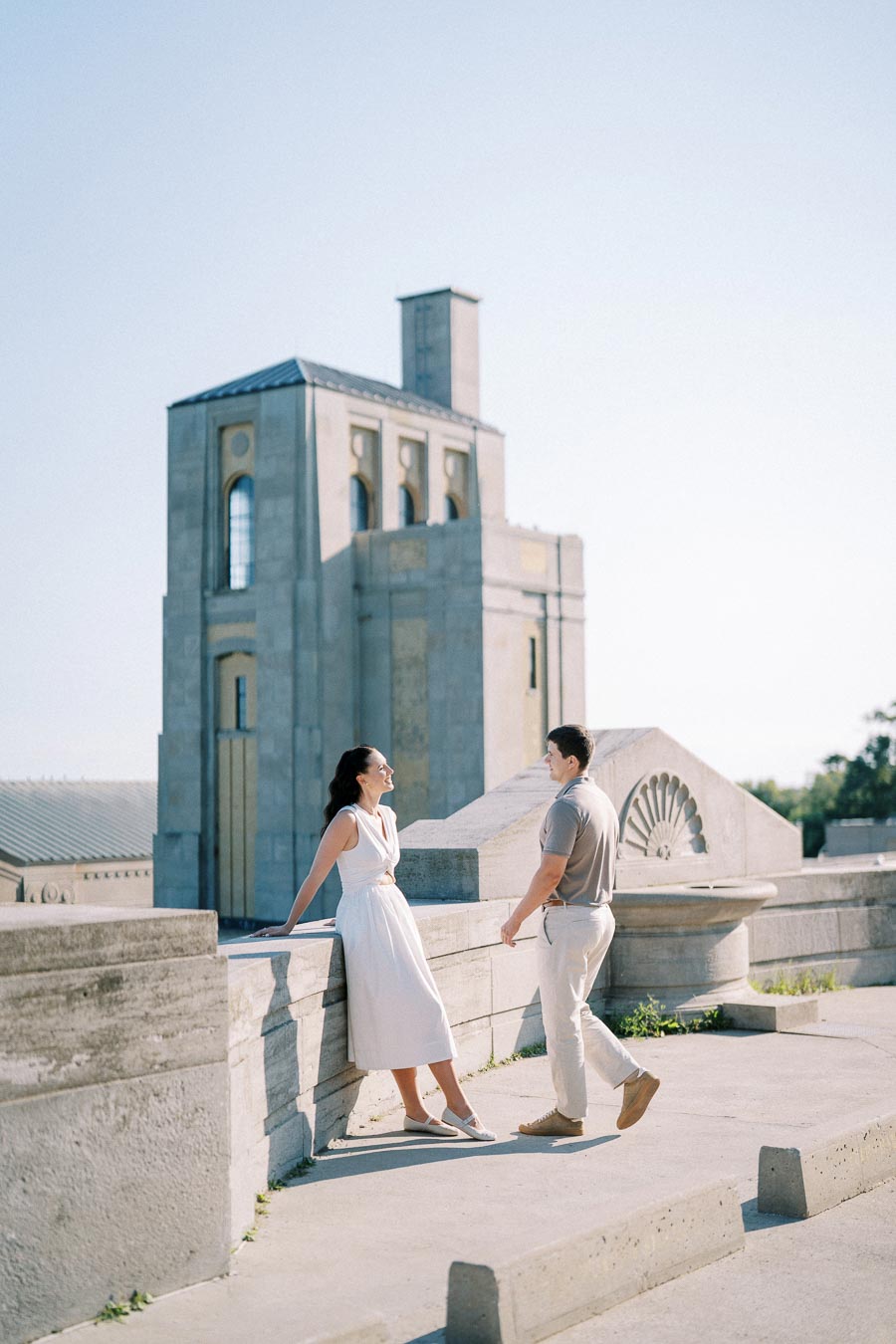 A woman in a white dress leans against a stone railing, conversing with a man in light casual attire. They are outside in a sunlit area, with an architectural building featuring tall, rectangular shapes in the background.