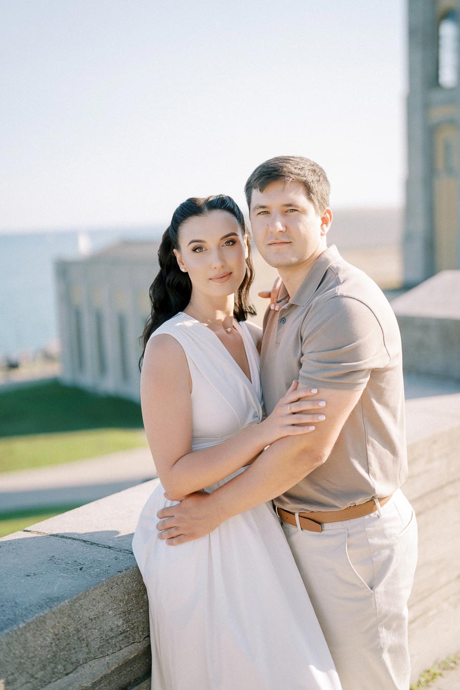 A couple embracing on a sunny day with a scenic ocean backdrop. The woman is wearing a white dress, and the man is in a beige polo shirt. They stand beside a stone railing.