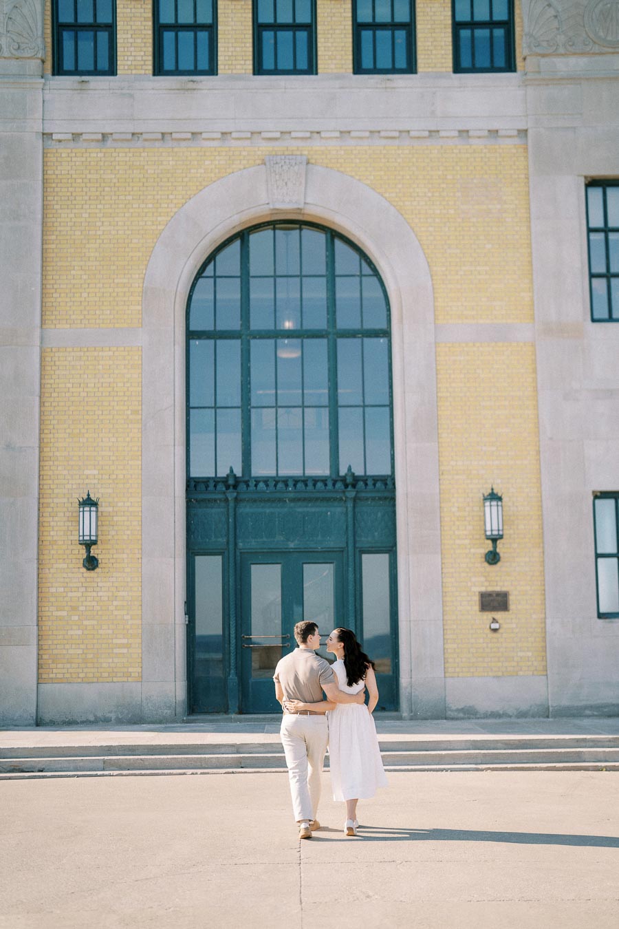 A couple walking hand in hand towards a large, arched entrance with yellow brick and tall windows, evoking feelings of romance and adventure.