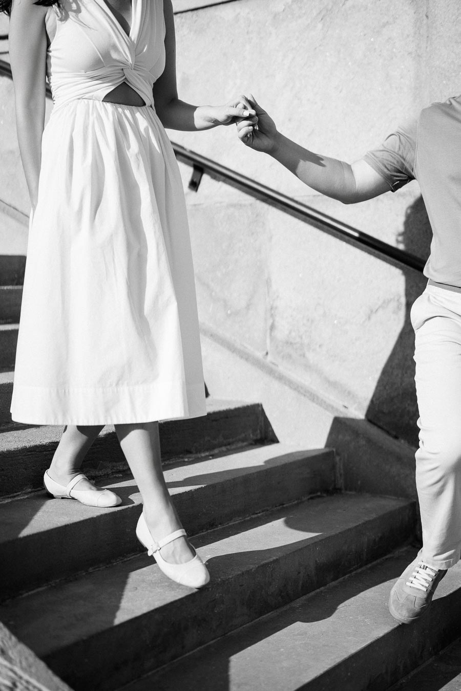A black and white photo of a couple holding hands while walking down a set of stairs. The woman is wearing a white dress and flat shoes, while the other person is wearing casual pants and sneakers.