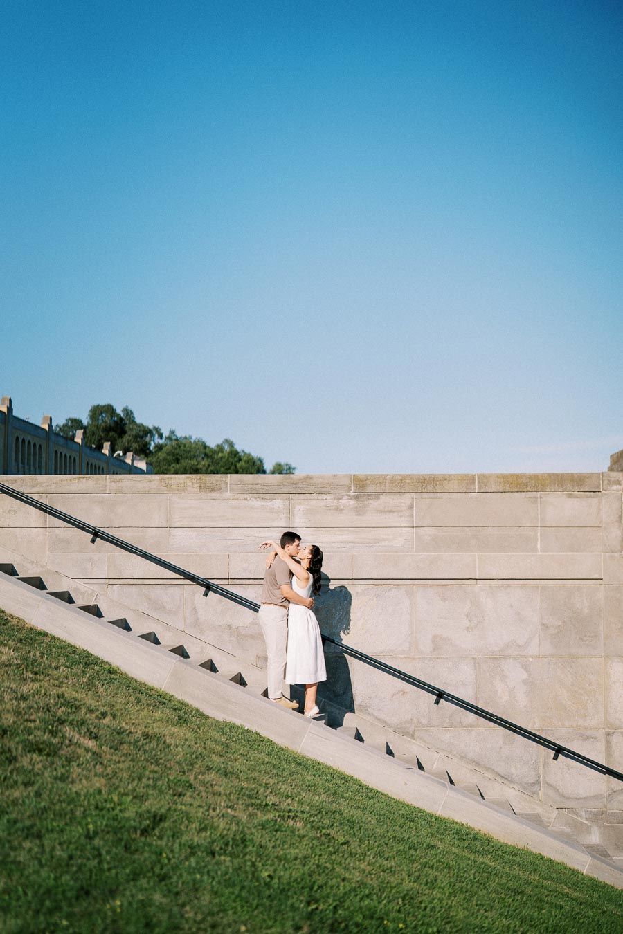 A couple embracing on a stone staircase with a clear blue sky, surrounded by greenery and architectural elements.