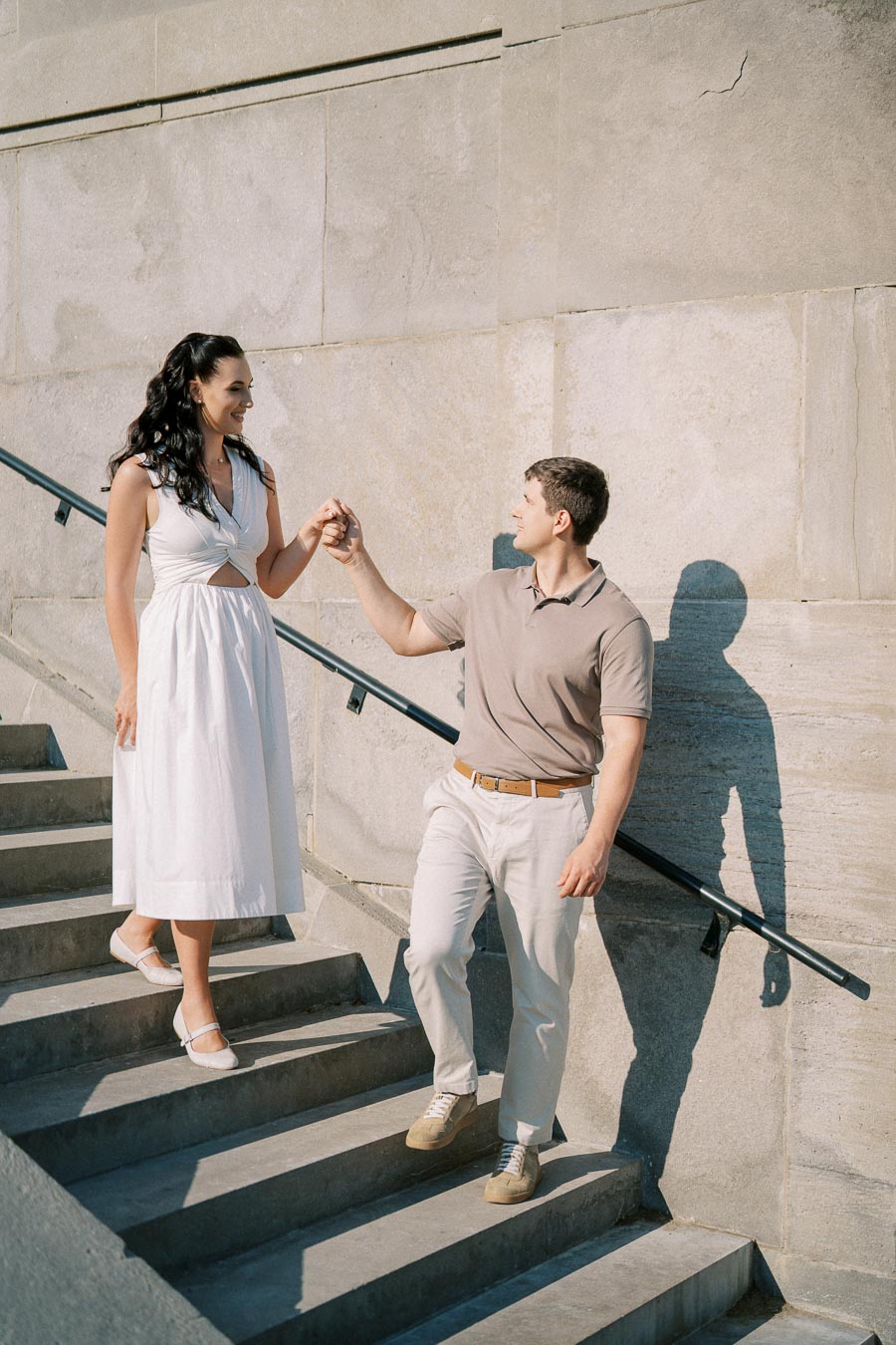 Couple walking hand in hand on stone steps, with a woman in a white dress and a man in a beige shirt, in front of a textured concrete wall.