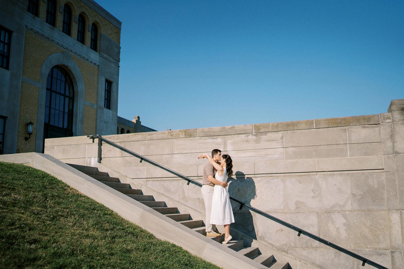 Couple embracing on outdoor stone staircase with historic building under clear blue sky.