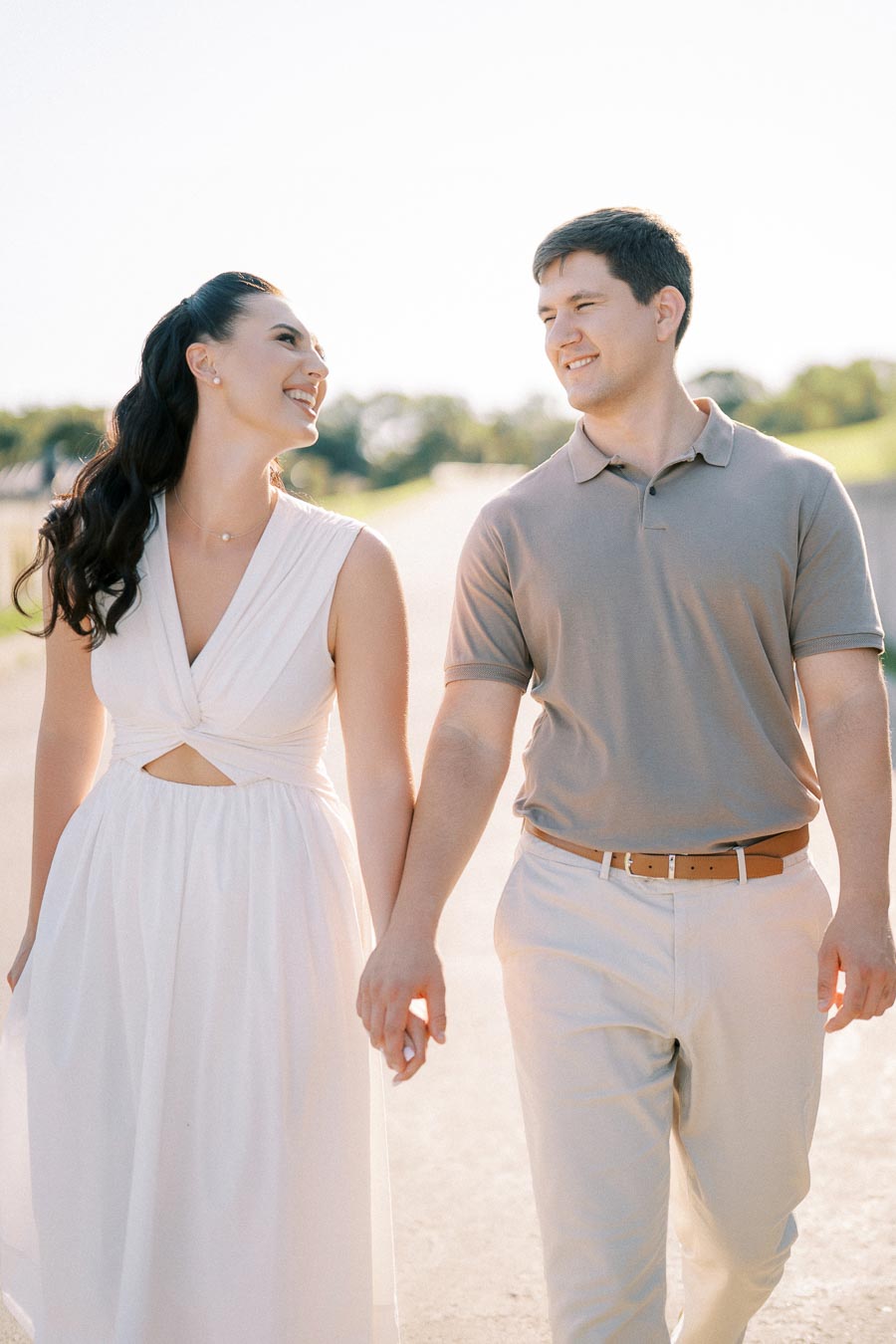 A smiling couple walks hand in hand on a sunny day, with the woman wearing a white dress and the man in a beige polo and pants, captured with a blurred natural background.