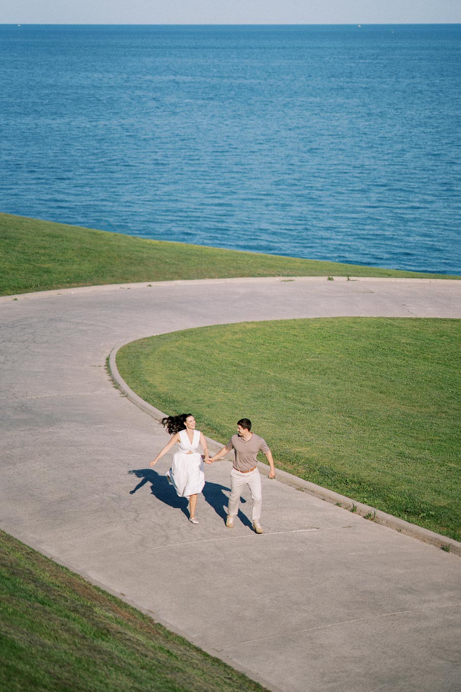 A couple joyfully runs hand in hand on a curved pathway by a scenic blue lake, surrounded by lush green grass under a clear blue sky.