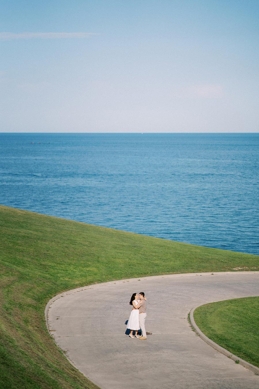 A couple embraces on a curved concrete path surrounded by green grass, with a vast blue sea in the background under a clear sky.