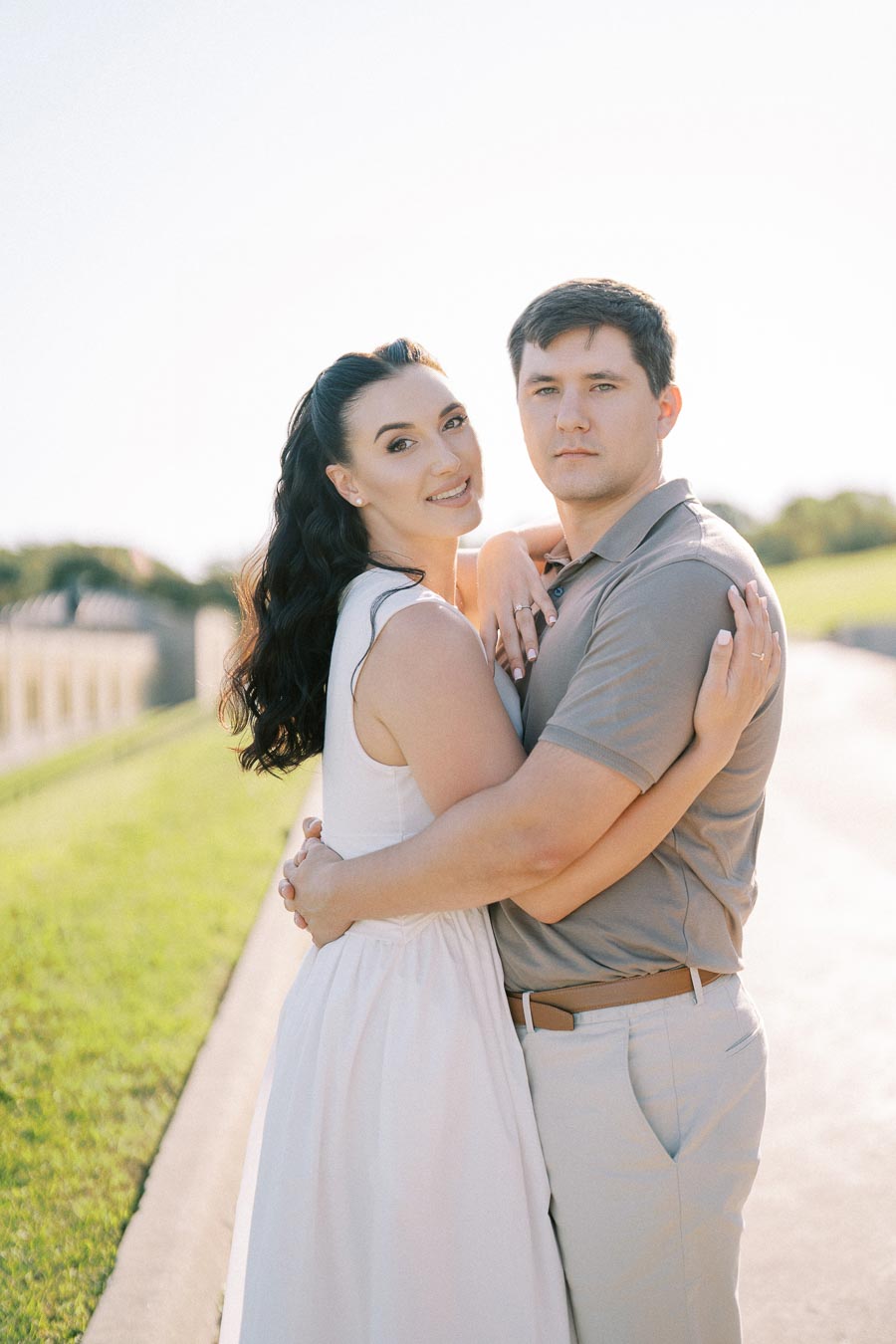 A couple embracing outdoors on a sunny day, with a lush green grass background and clear blue sky. They are smiling and dressed in light, casual attire, highlighting a serene and joyful moment.