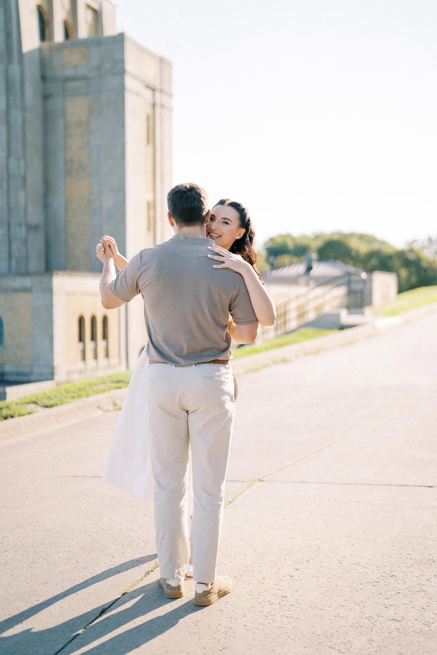 A couple dancing outdoors in the sunlight, with a historical building in the background. The man is wearing a gray shirt and light pants, while the woman embraces him, smiling. The scene conveys a romantic and joyful moment.