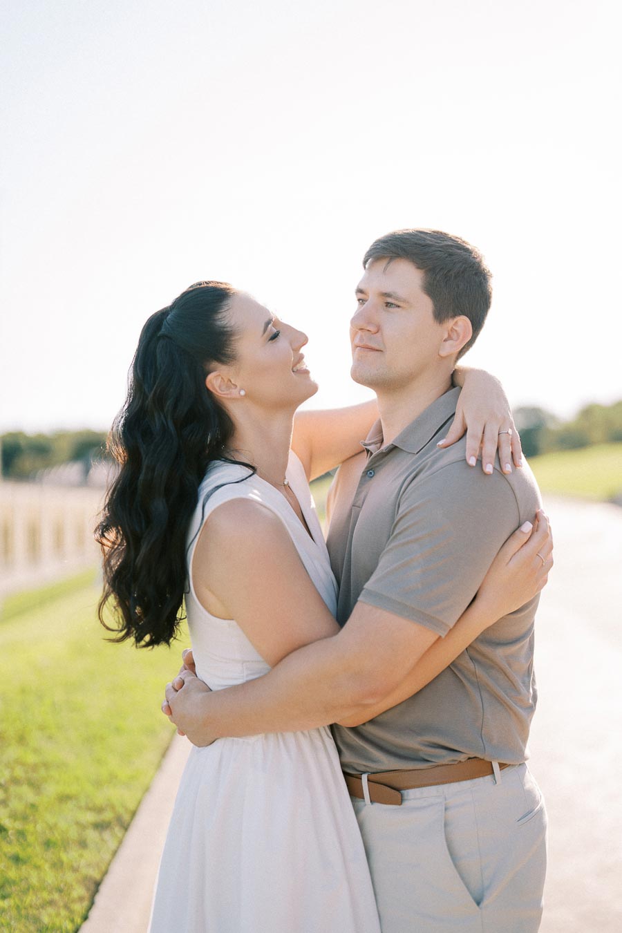 A couple embracing under a clear sky, standing on a sunlit path with green grass in the background, conveying a romantic and joyful moment.