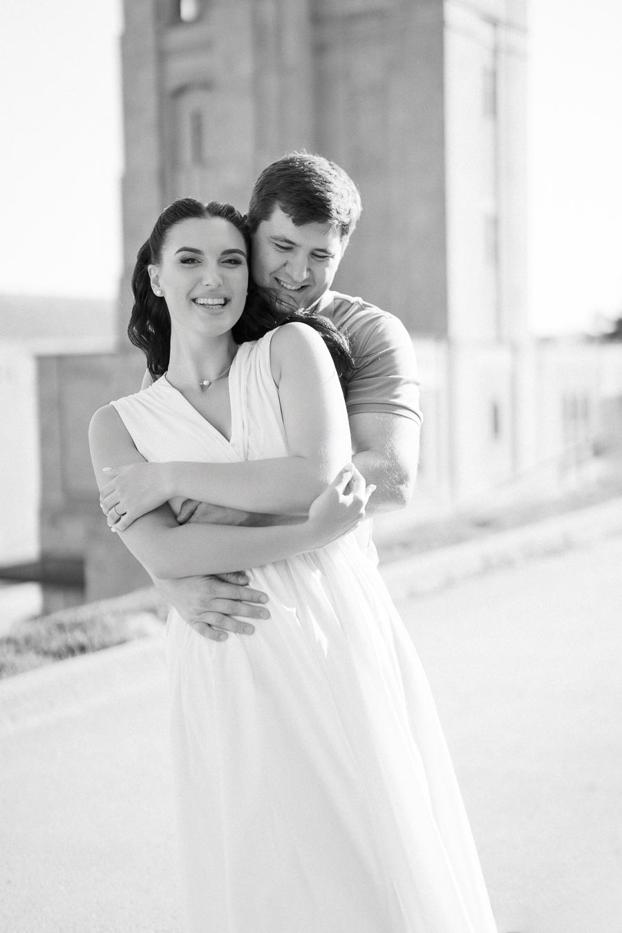 A happy couple embracing outdoors in black and white, with a blurred building in the background, depicting love and romance.