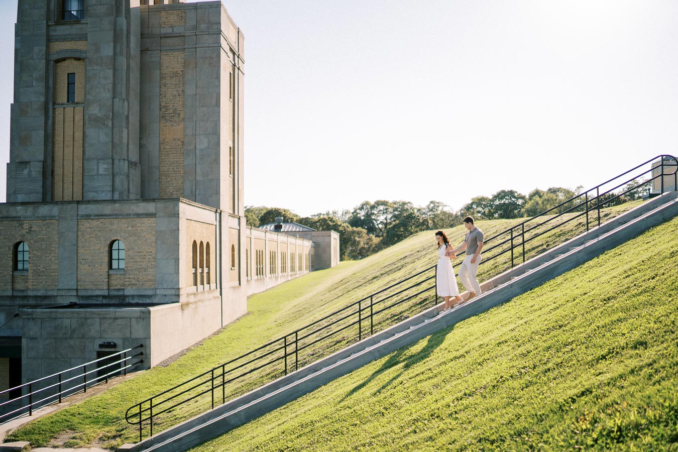 Couple walking down staircase beside historic stone building on a sunny day, with green grassy slope and blue sky in the background.