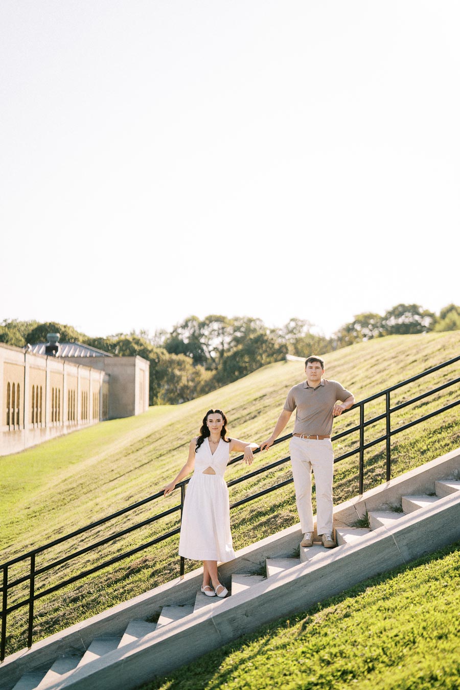 Couple standing on outdoor stairs with green grass and a building in the background under a clear sky.