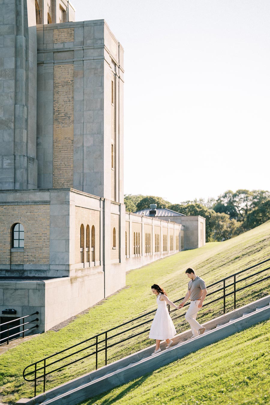 A couple walking down a set of outdoor steps alongside a historic building with arched windows, surrounded by a sunny green landscape.