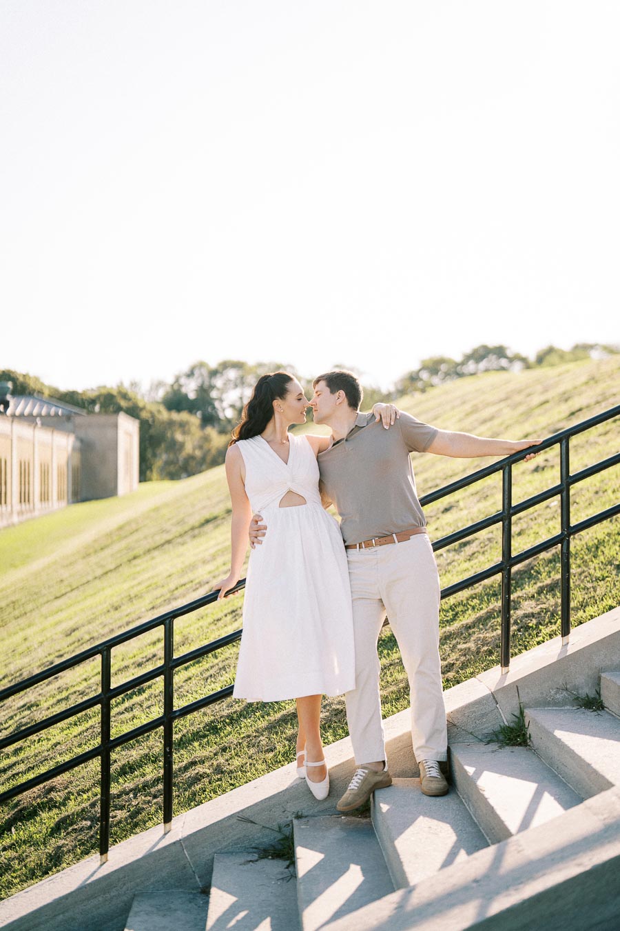 A couple standing on outdoor stairs, embracing and leaning in for a kiss, with a bright sky and a lush green hillside in the background.
