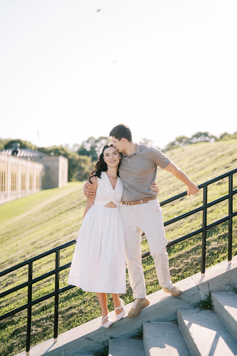 A couple embracing on outdoor steps with a scenic grassy hill and a building in the background, under a clear blue sky.