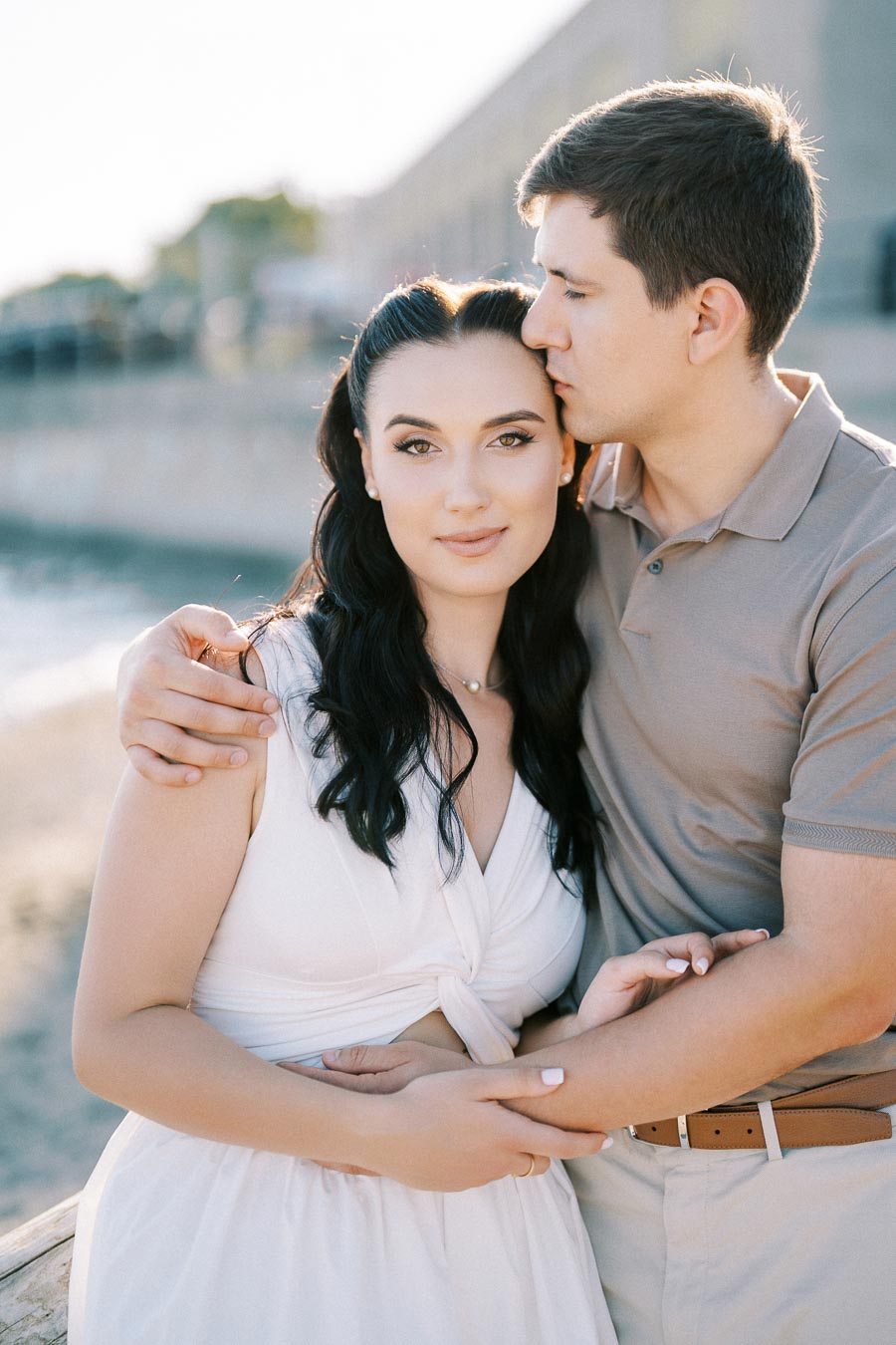 A romantic couple embracing on a sunny beach, with the woman in a white dress and the man in a beige shirt, highlighting love and serenity.