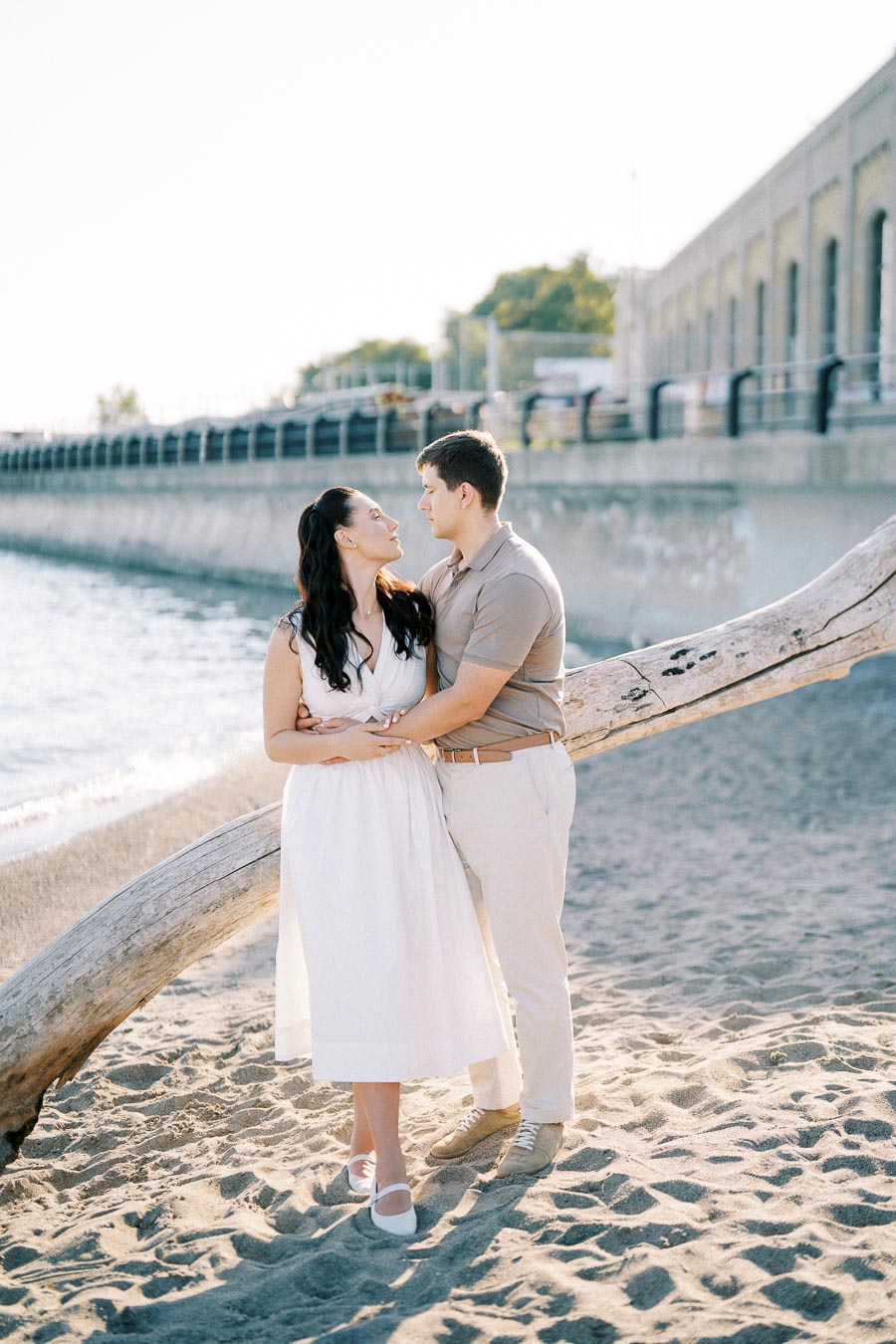 Romantic couple embracing on a sandy beach by the water, with a driftwood log and a stone wall in the background.