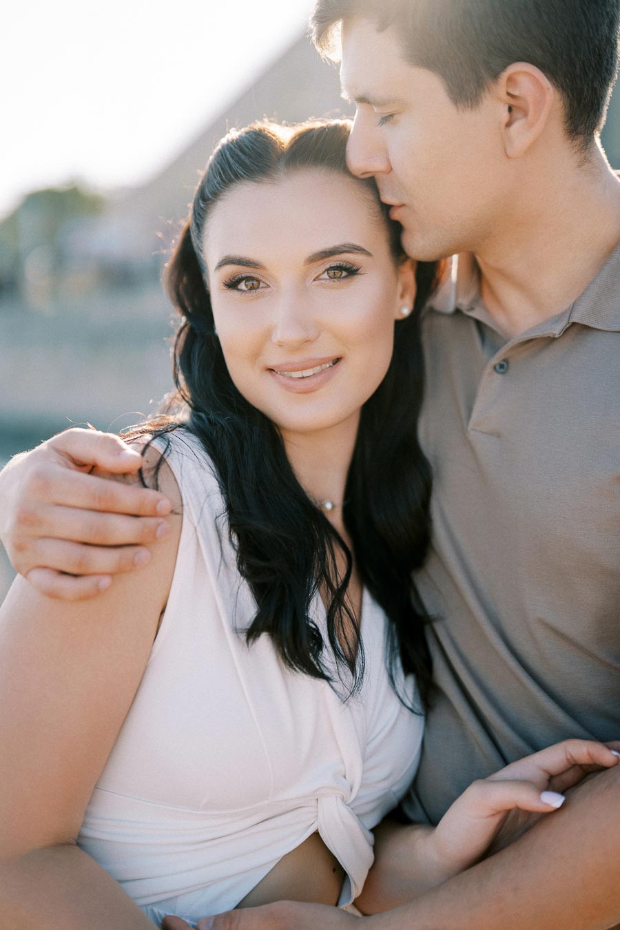 A couple embraces lovingly, the woman smiling warmly at the camera and the man gently kissing her forehead, in a sunlit outdoor setting.