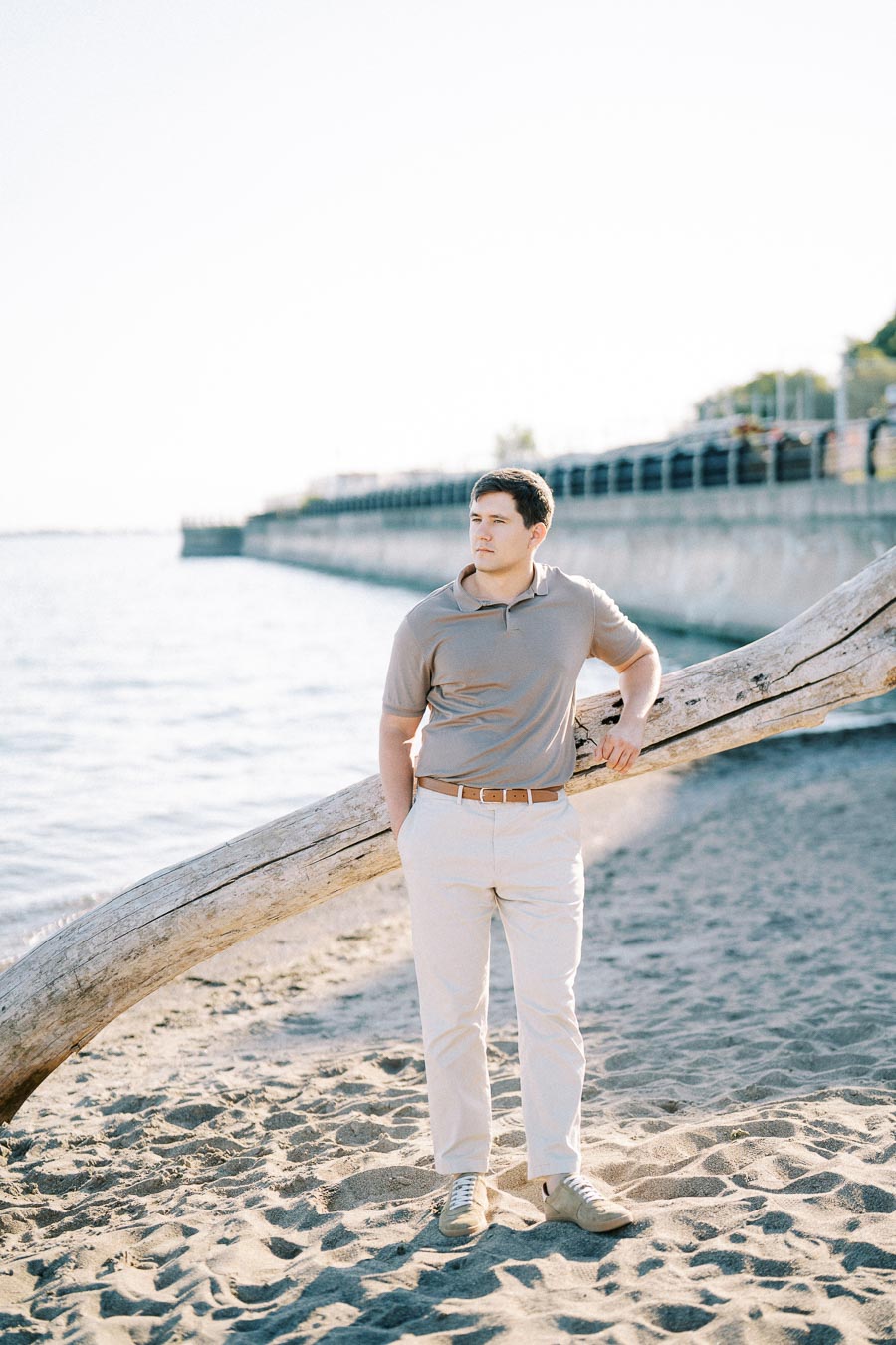 Man standing on sandy beach next to a large driftwood log, wearing a beige shirt and pants, with a scenic waterfront and boardwalk in the background.