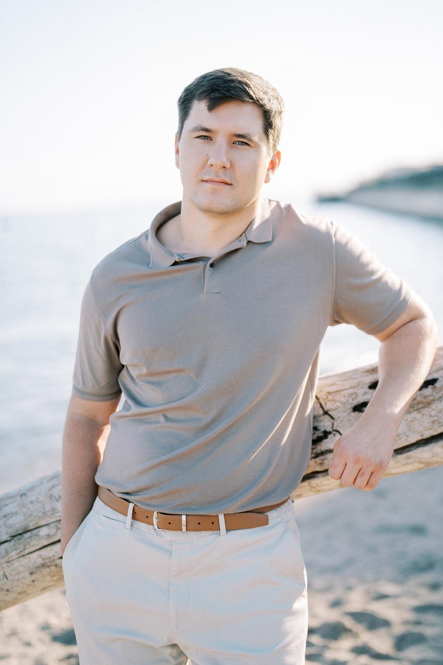 Young man in a beige polo shirt standing by a driftwood log on a sunny beach with calm ocean in the background