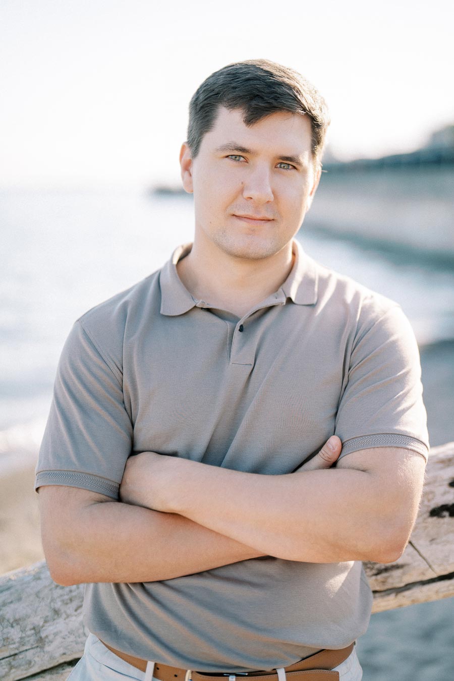 Person in a gray polo shirt standing by the beach with arms crossed, smiling slightly, with a blurred ocean background.