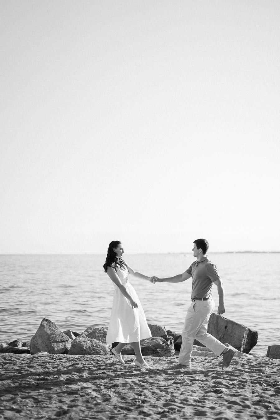 A couple holding hands and smiling while walking on a rocky beach by the ocean, captured in black and white.