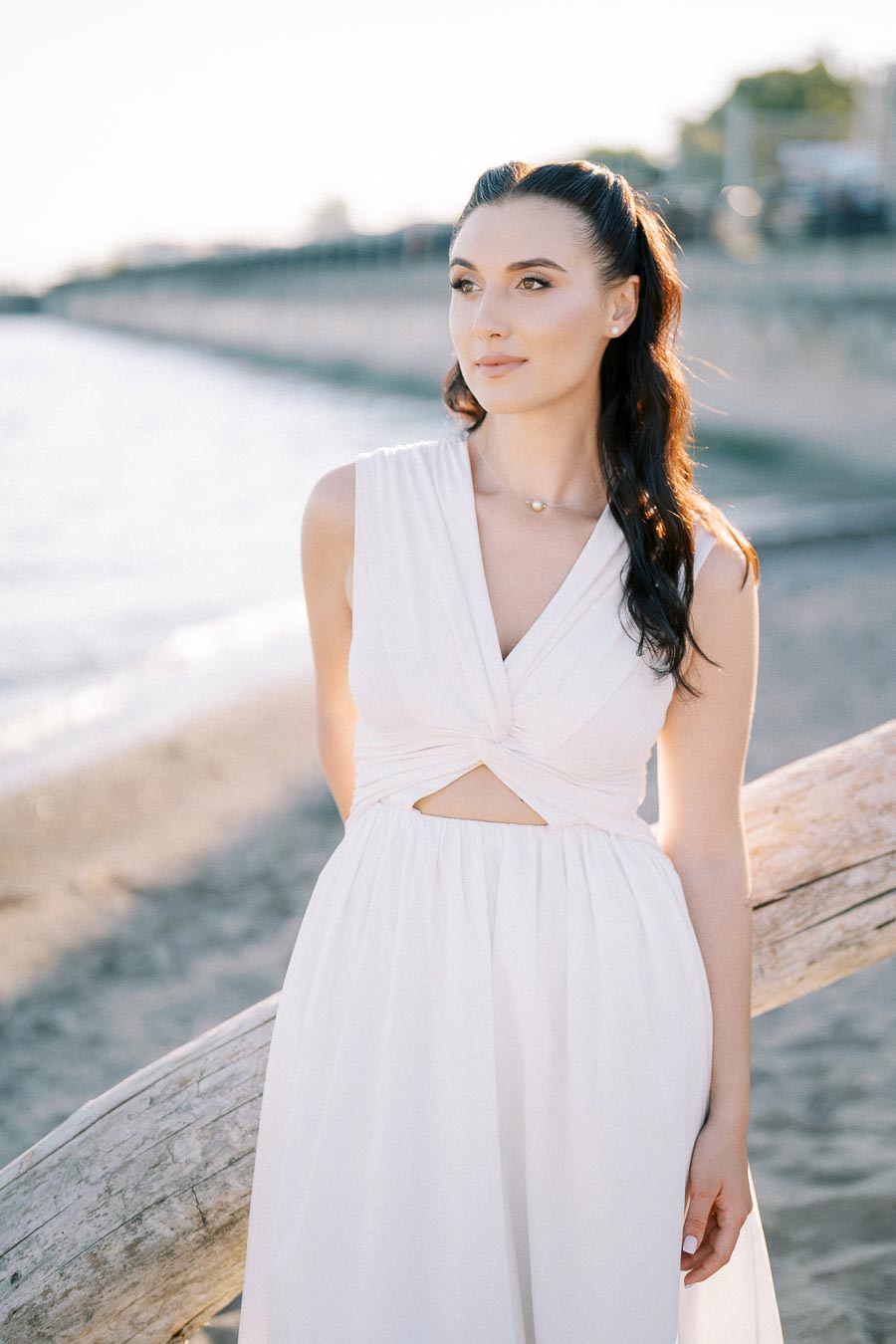 A woman in a white dress stands by a beach, leaning on a wooden railing, with the ocean and a sunny sky in the background.