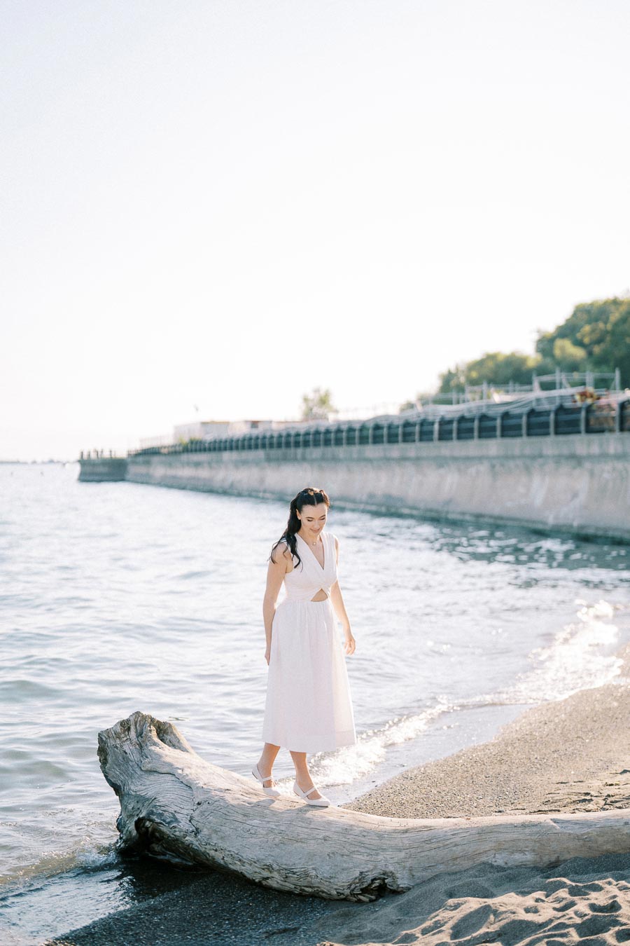 A woman in a white dress walks on a large driftwood log at a beach with gentle waves and a distant boardwalk under a clear sky.