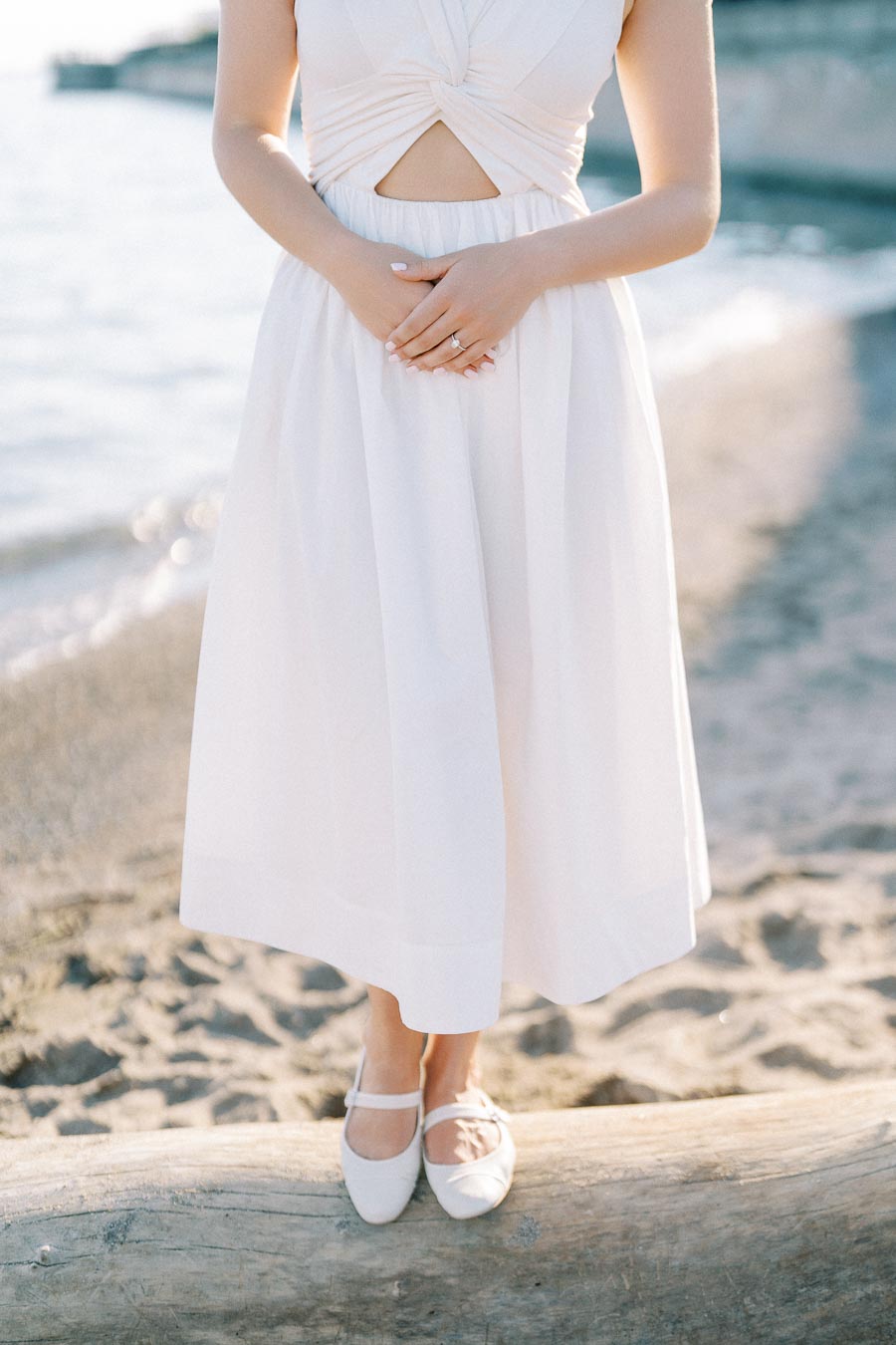 A woman in a white summer dress and shoes stands on a driftwood log at a beach, with softly blurred ocean waves in the background.