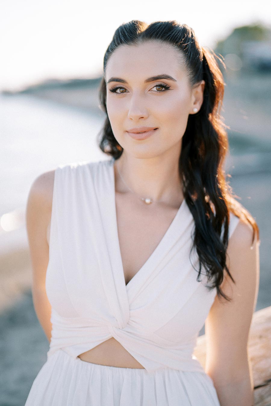 A person with long dark hair wearing a white dress standing by the beach, with a soft-focus background.