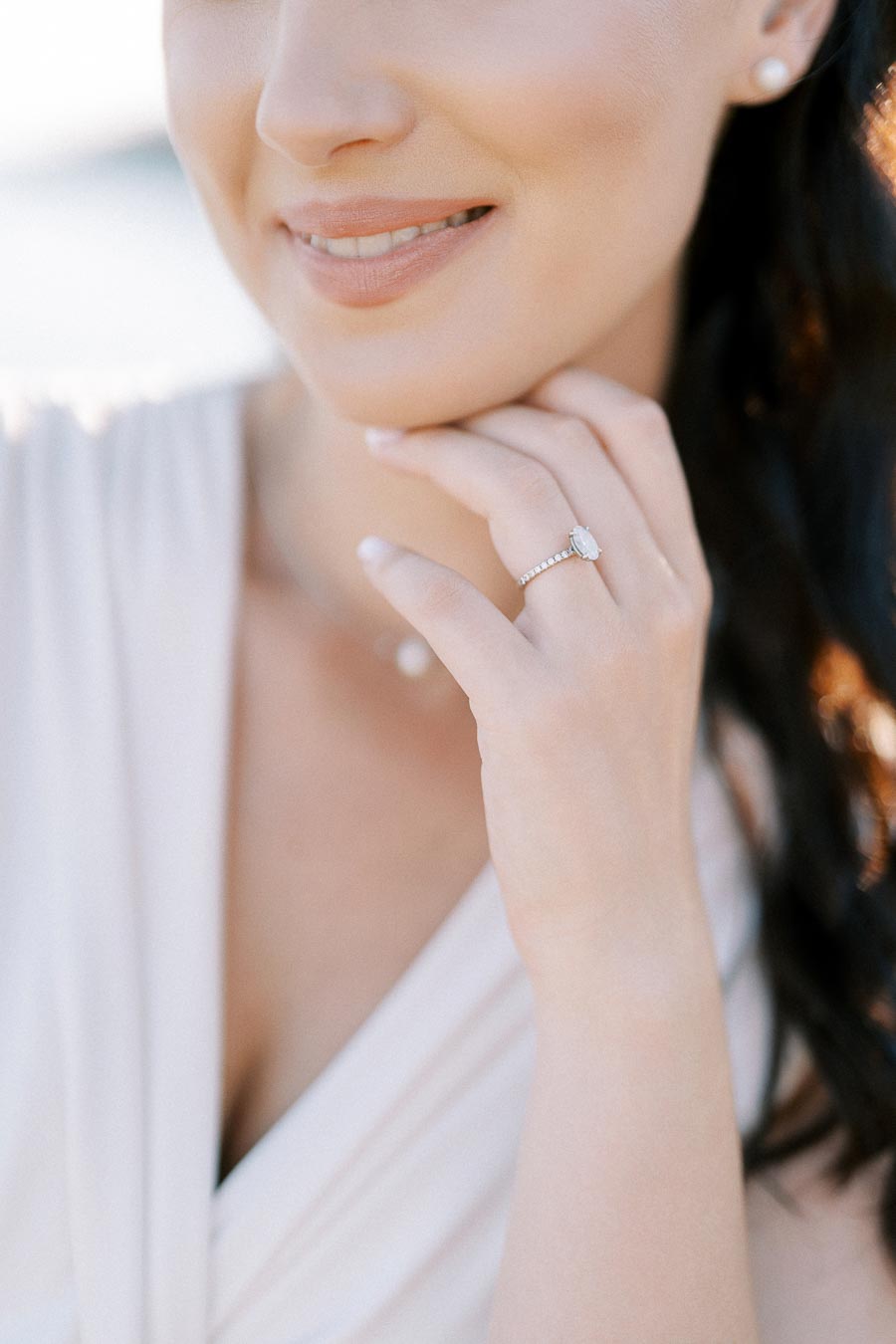 Smiling woman wearing an elegant diamond engagement ring and pearl earrings, gently touching her chin with a serene backdrop.
