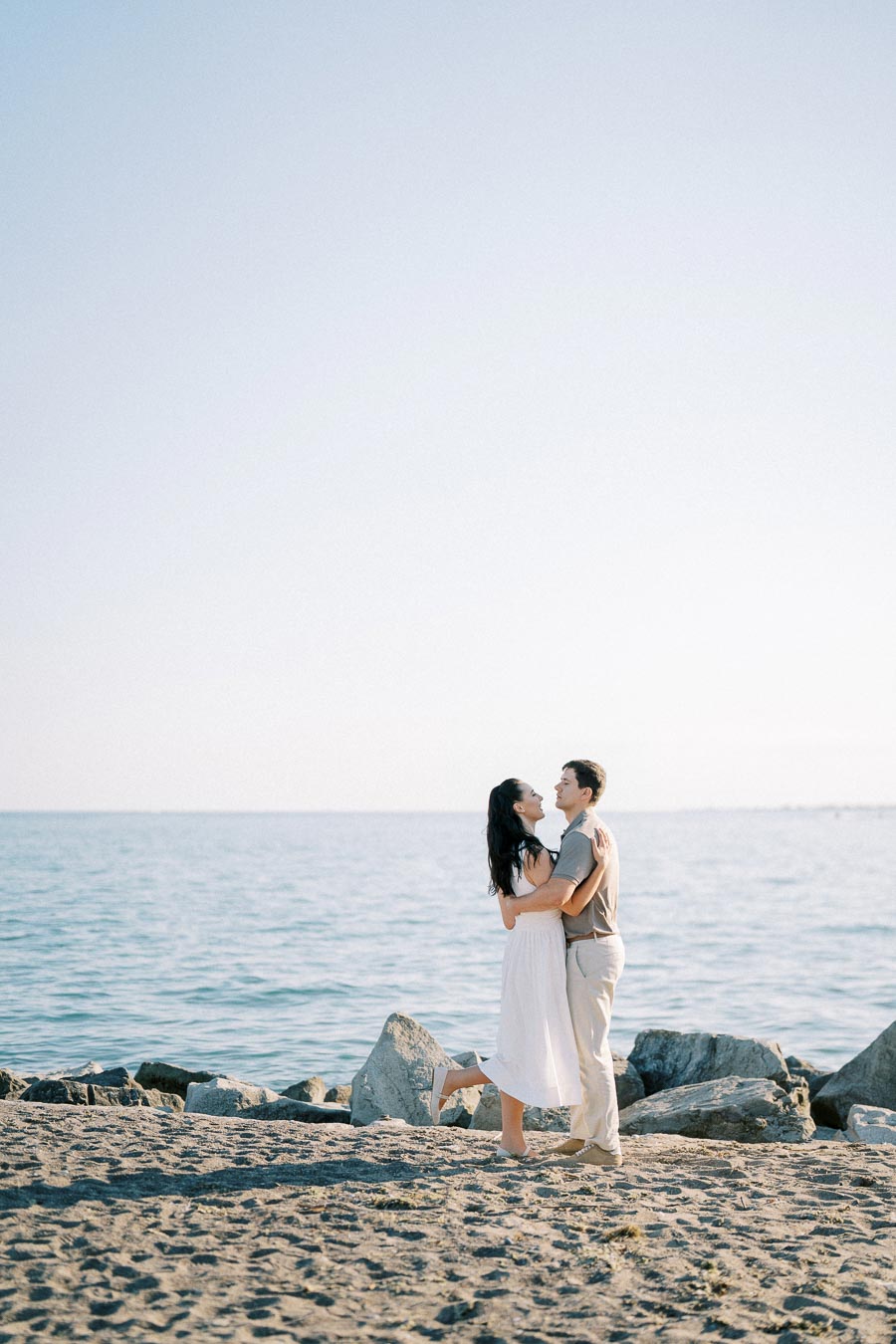 Romantic couple embracing on a sandy beach by the ocean, under a clear blue sky.